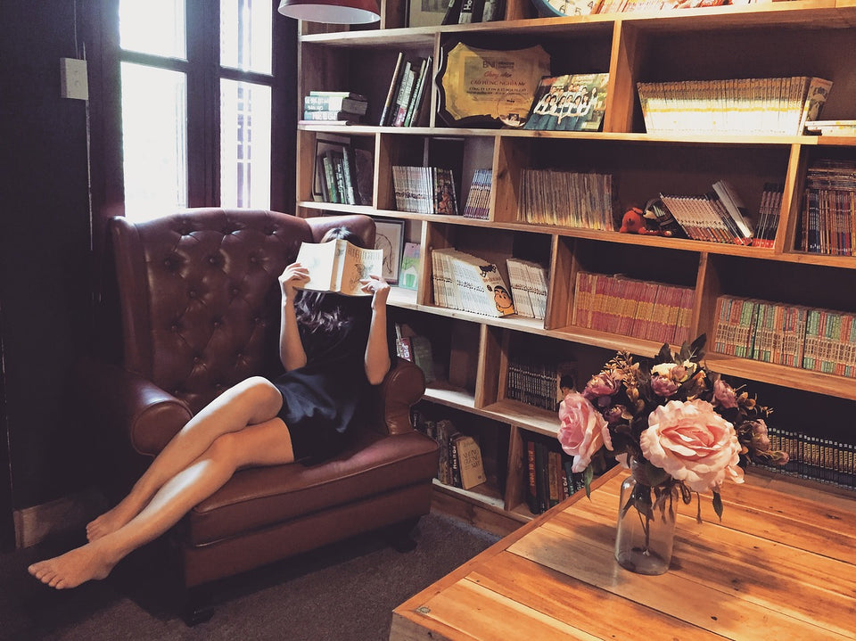 woman reading on couch in front of bookshelf lined with books. Coffee table, vase with roses